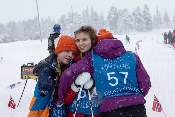 Tre glade barn i vinterklær som klemmer hverandre under et skirenn i snøvær. To av barna har på seg blå startnummervester hvor det står «Barnas Ridderrenn».