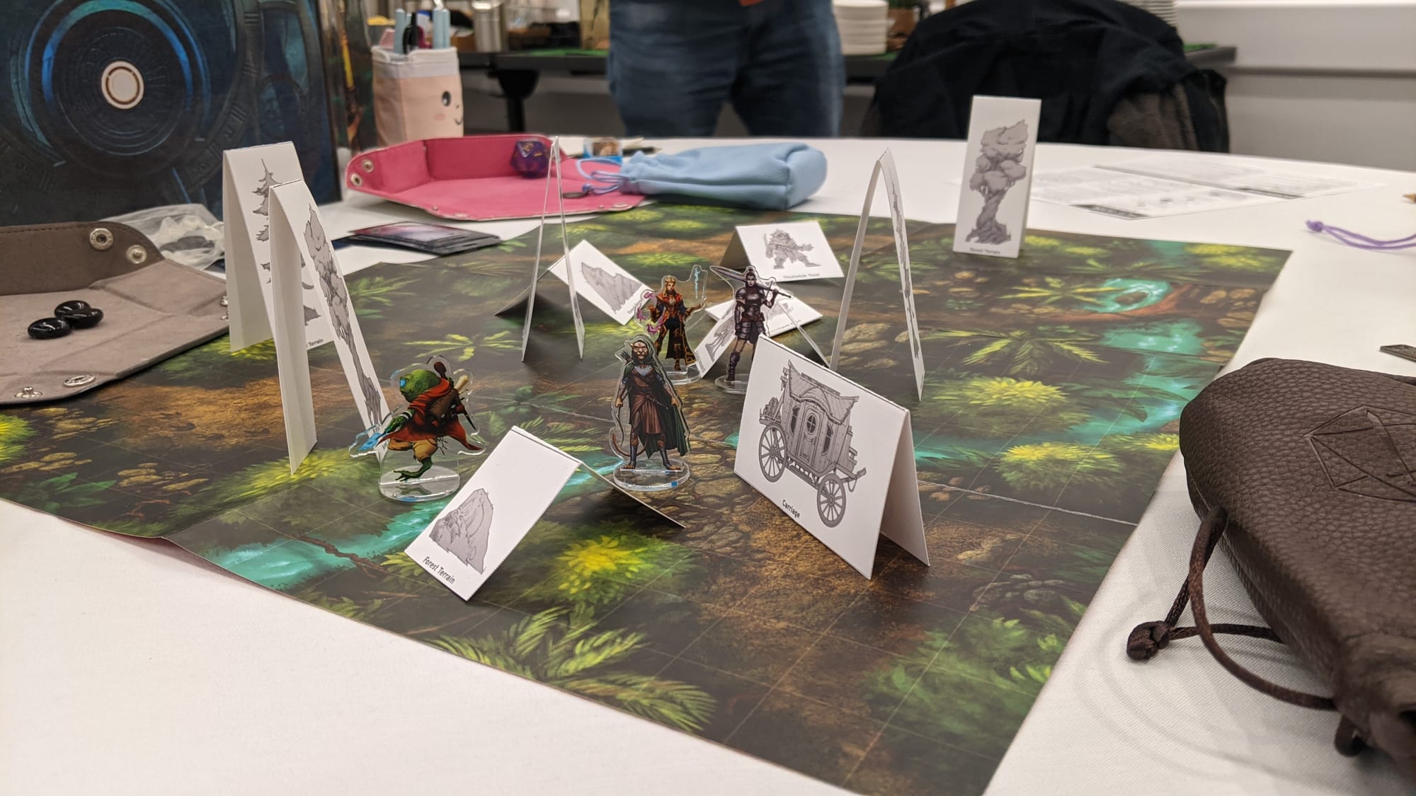 A white table festooned with folded paper miniatures, plastic character figures and a forested battle map.