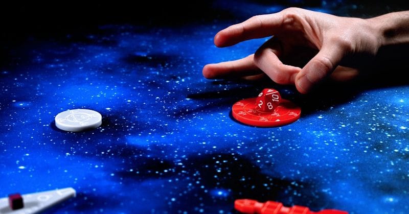 Hand poised to flick a red die. Blue and black stellar playmat; red and white pieces.