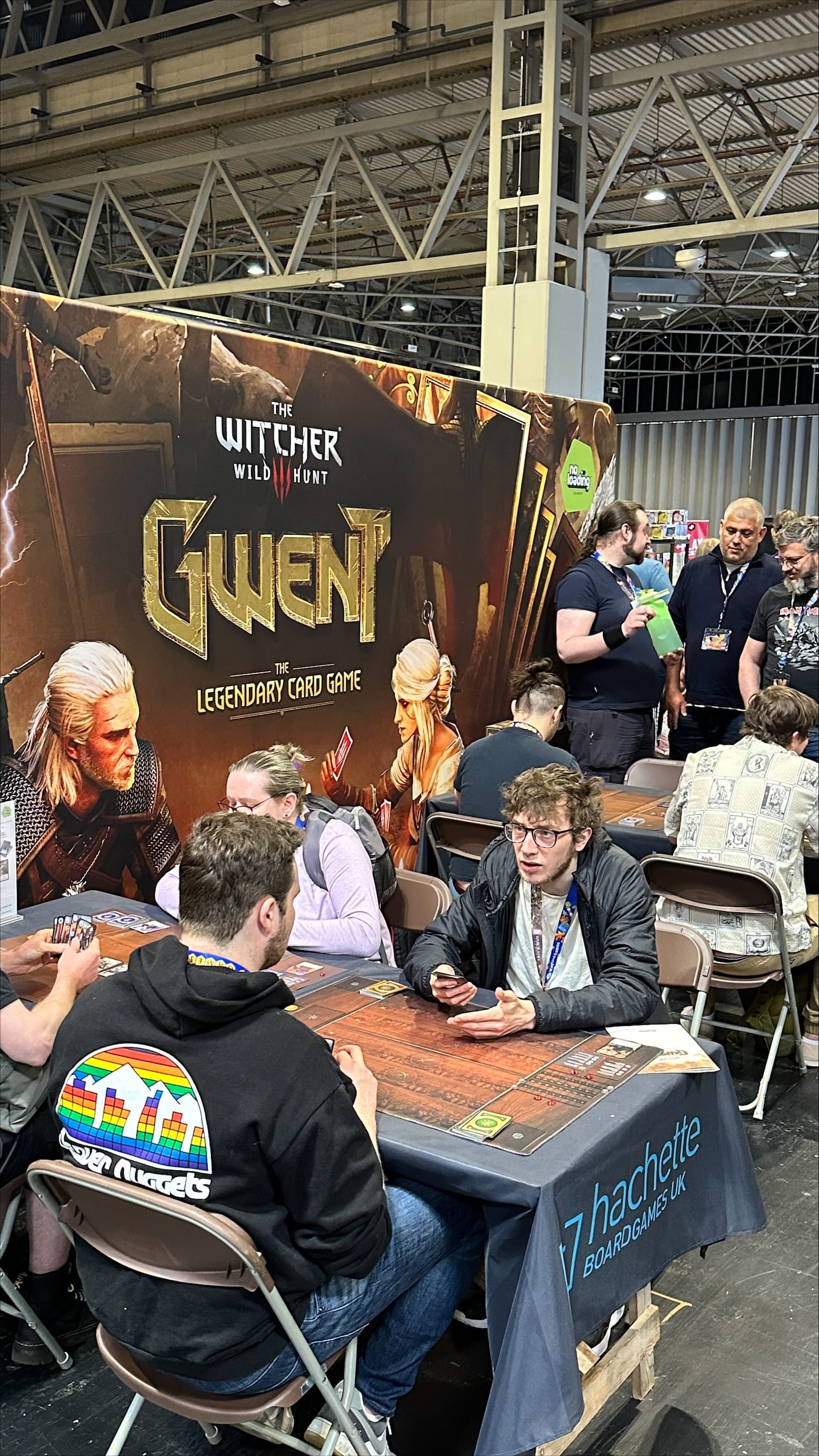 Six people sitting at folding tables playing the Gwent card game. Promotional poster for the game flanks them.