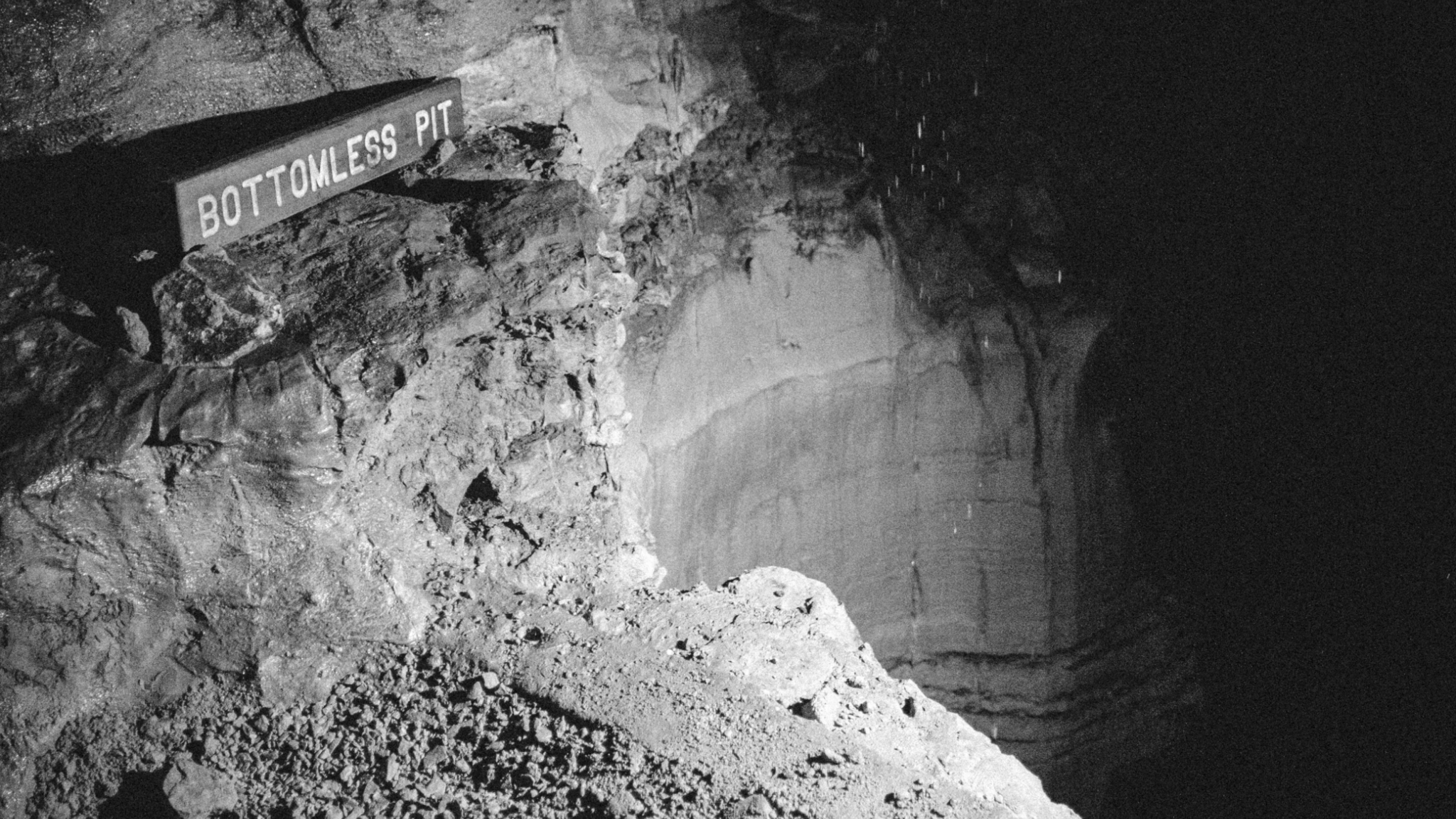 A photograph in black and white of a pit descending into darkness. There's a sign labelled "BOTTOMLESS PIT". 