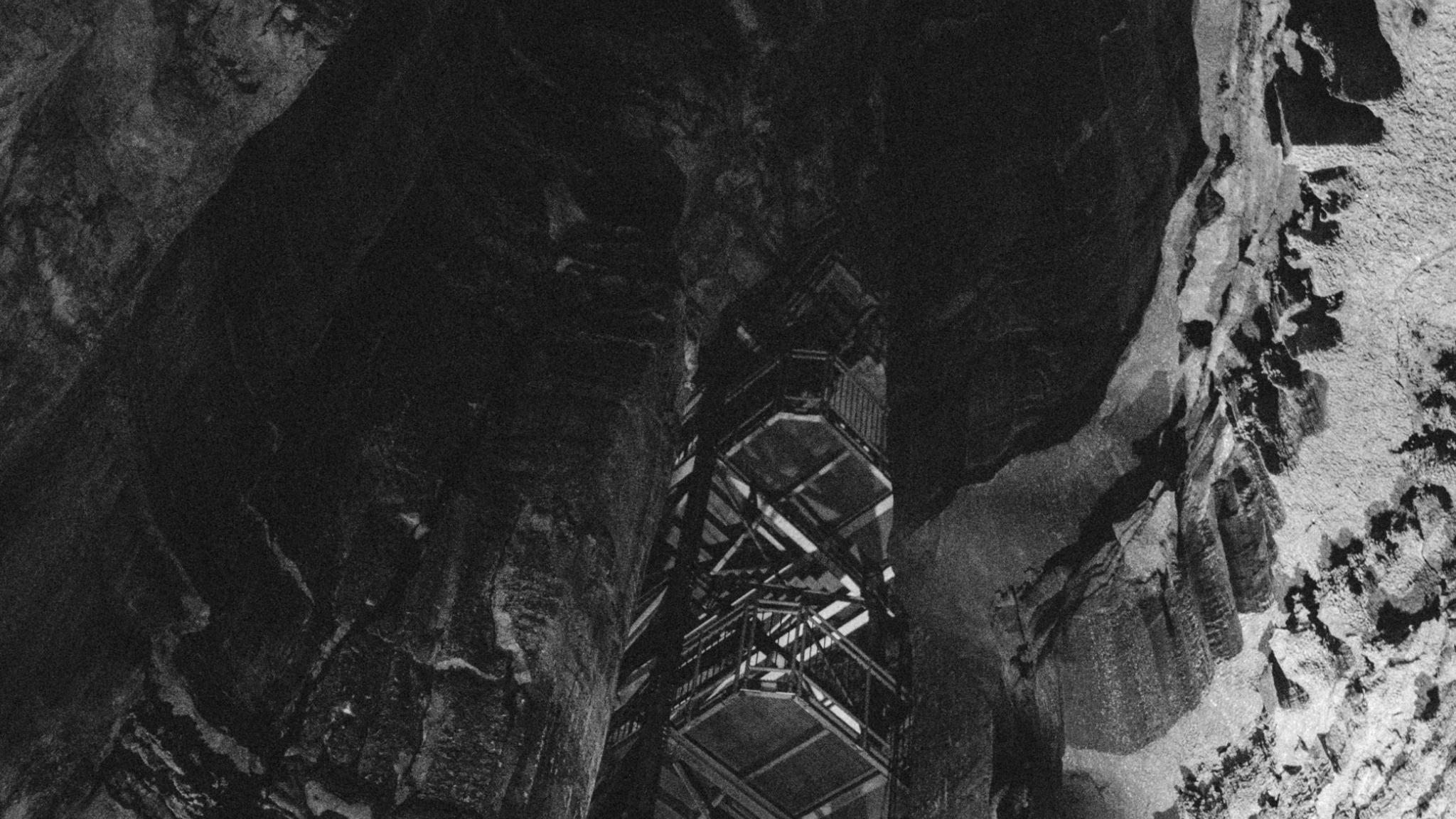 A photograph in black and white of the inside of a cave, looking up at the chamber ceiling. There's a built structure in the cave, with odd scaffolding. With the stark lighting, it almost looks like there are stairs moving up the chamber.  