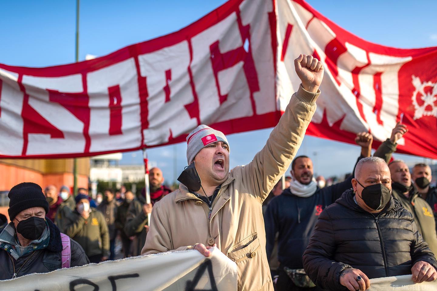 A protest photo. A man raises a fist and shouts. A crowd is in the background, holding a red and white banner.