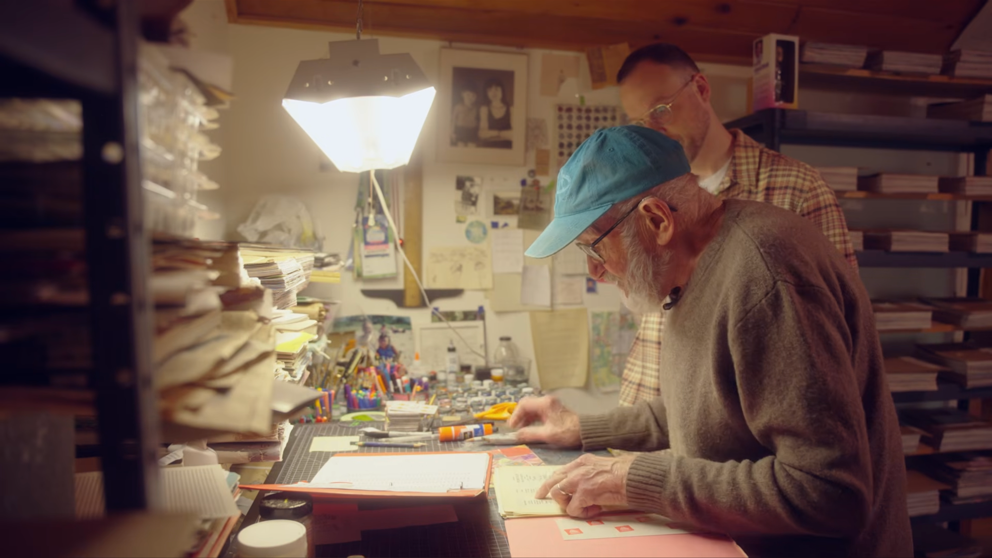 Two men, one in a blue cap, stand in a cozy artstation full of shelves, paint pots, brushes, and paper. A bright workshop light hangs from the top.