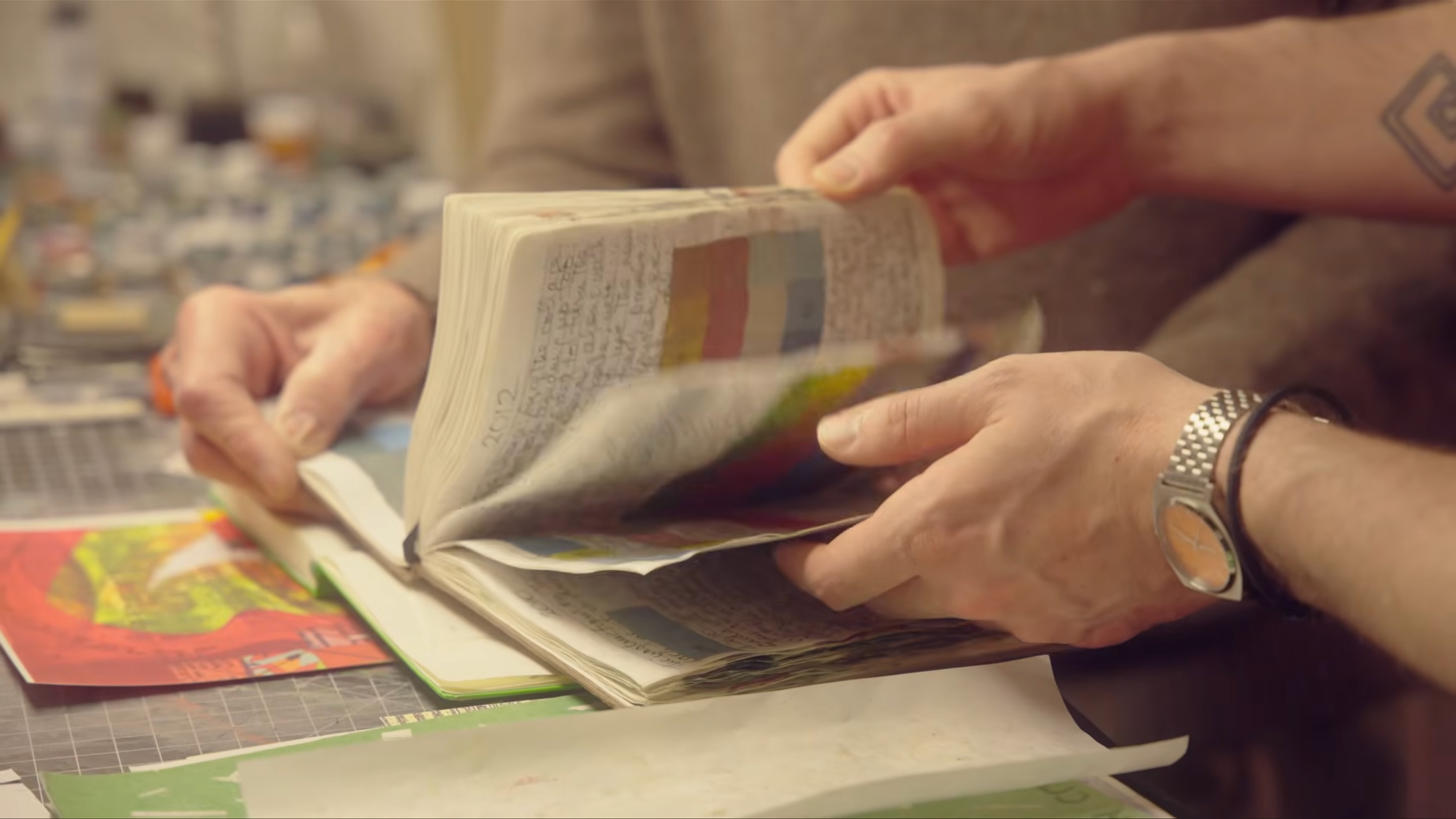 Two people hold an old journal open, revealing handwritten text and colored swatches across every page. We see just their hands, the bottom foreground one wearing a silver watch.