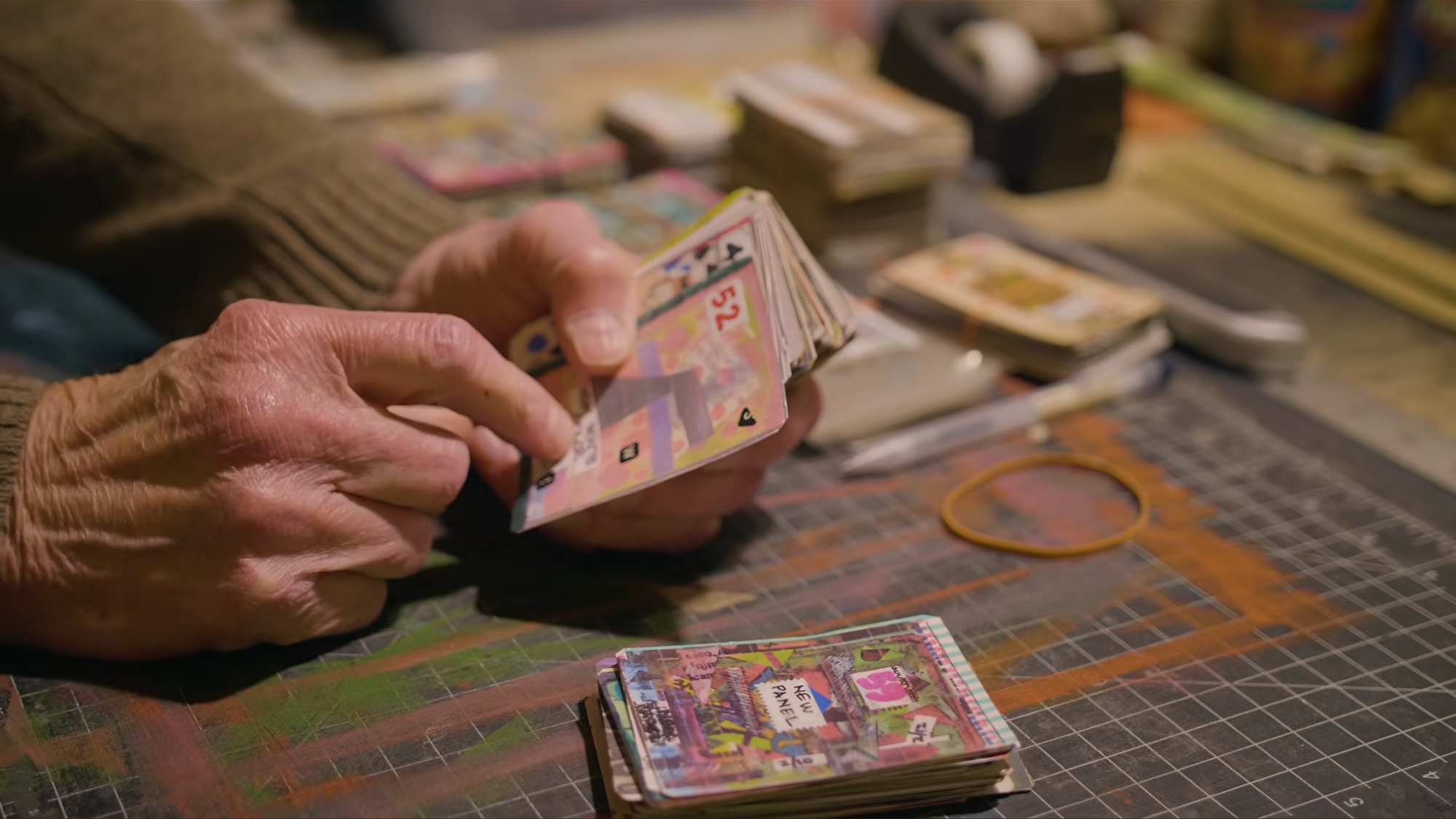 An elderly man flips through a deck of motley, collaged cards on a well-loved art station.