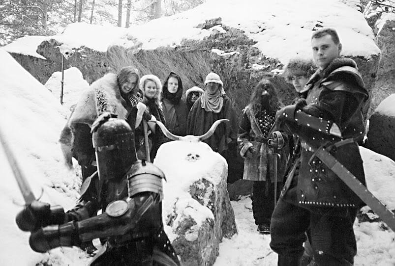 Nine people posing in medieval-inspired cosplay gear against a snowy forest backdrop. A skull and cow horns adorn a boulder in the middle.