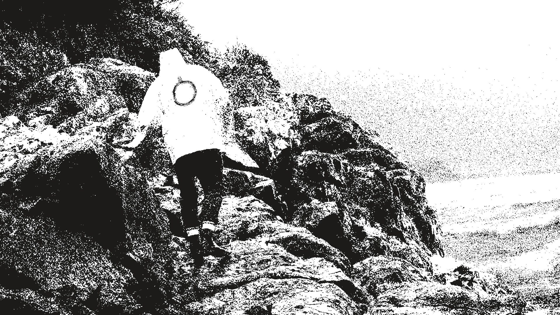Midnight of the Century. Evocative black and white. A lone figure scrambles over a rocky shoreline, wearing a light-coloured coat with the Oroboro logo on it, a circle with a tick at the midnight position.
