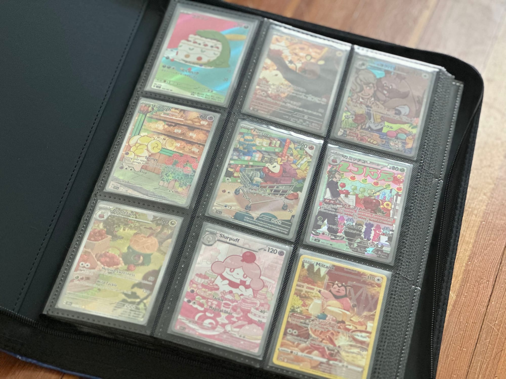 A black binder full of full art Pokémon cards sitting open to its first page on a wooden table.