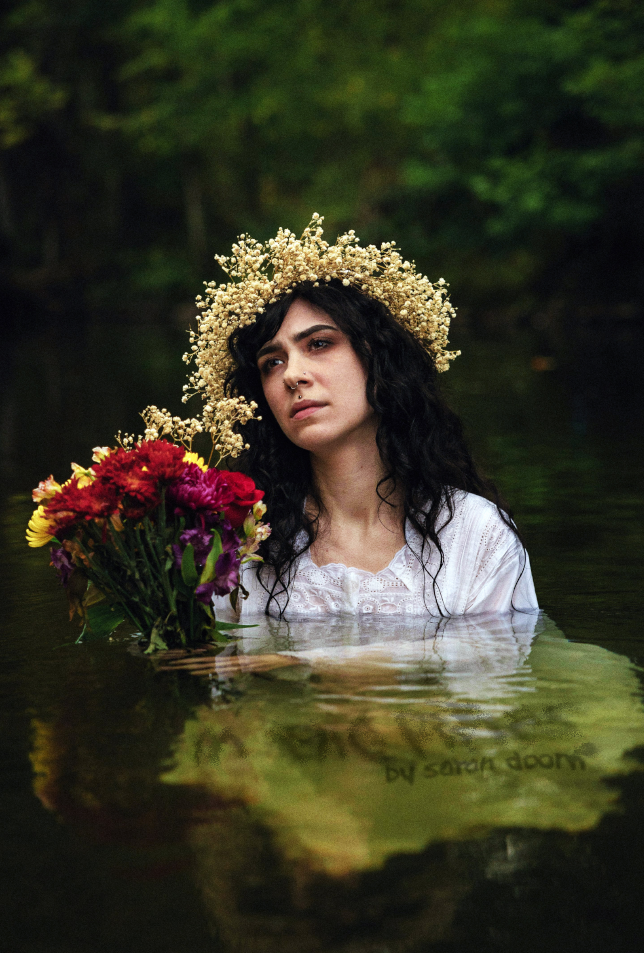 A person in a flower crown and white shirt holds a bouquet of flowers while standing in a body of water in the woods.