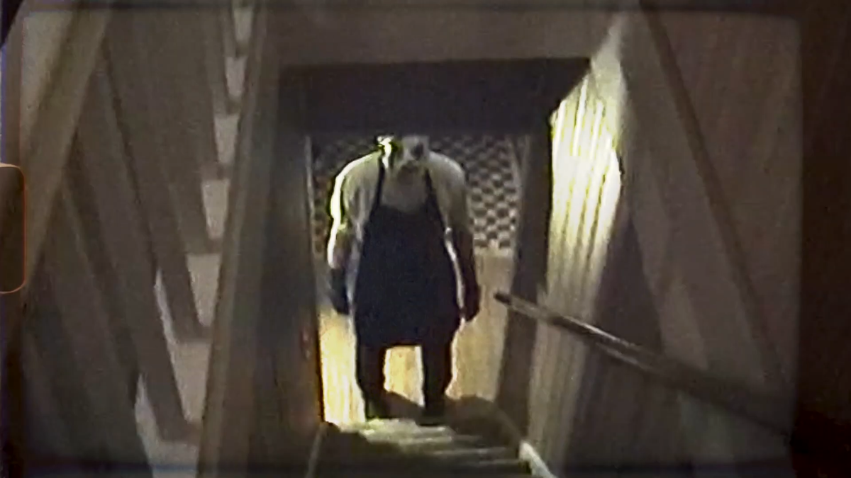 A person in a blck apron and deformed mask stands ominously at the bottom of a staircase.