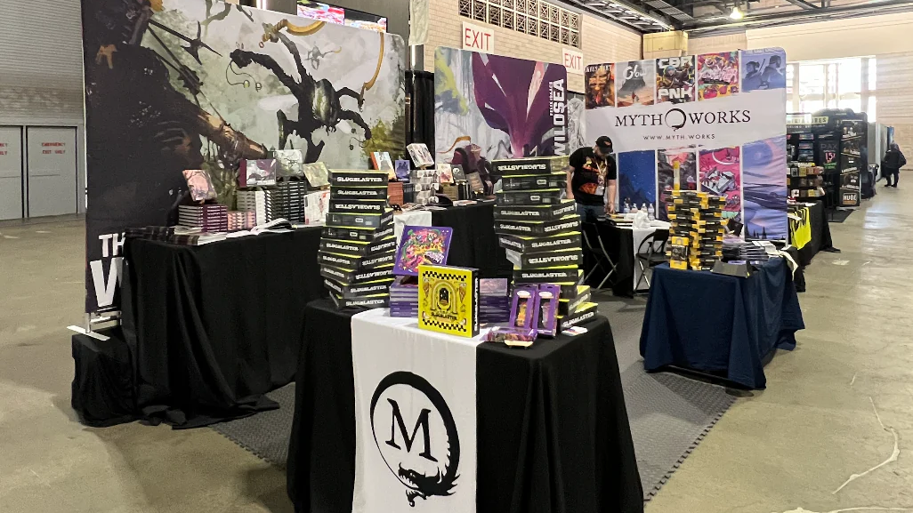 Tables covered in black and white cloths, RPG books and Mythworks branding. Three panel posters flank the small space.