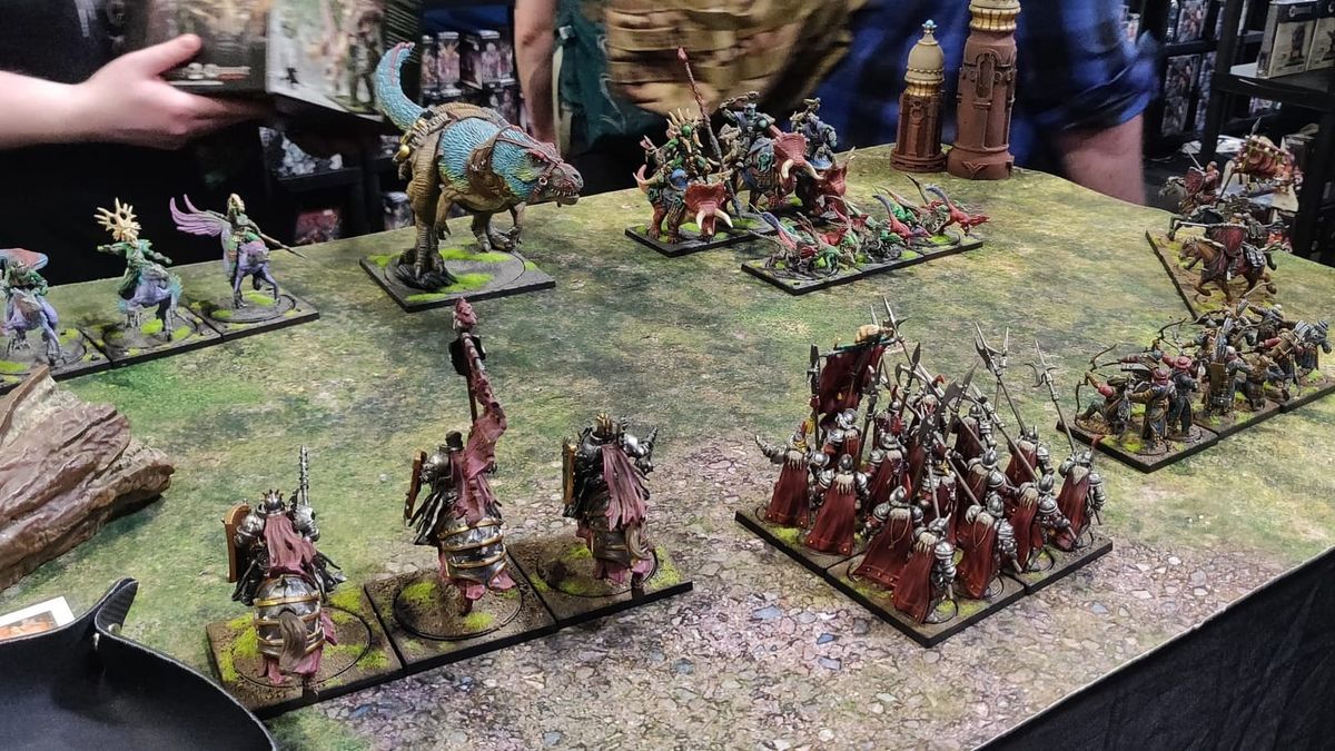 Two miniature armies on a green and brown table. One is knights in red capes, the other is mounted dinosaurs.