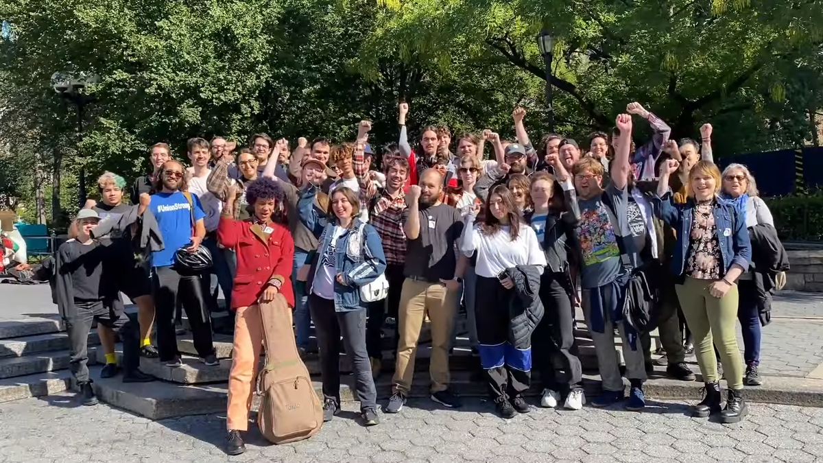 Dozens of people standing in a sunny pary with their fists raised in a gesture of solidarity. Wearing everyday clothing.