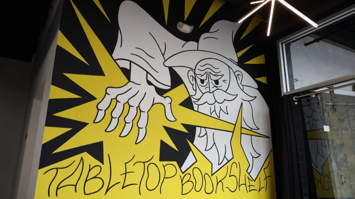 Concrete wall mural of a scowling wizard (black and white) casting a bright yellow spell with downturned hand. Stylized letters read: Tabletop Bookshelf.