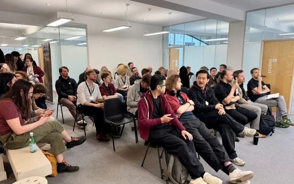 White, well-lit room full of people at a convention, most sitting in black chairs or on a bench. They face one direction and are chatting amongst themselves.