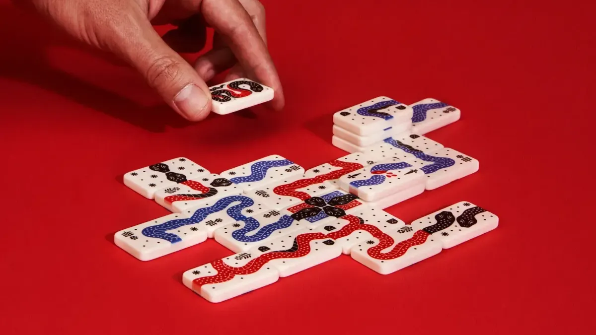 White game tiles featuring red, blue, and black snake illustrations on a red background. A hand reaches down from the top-left to place a tile.