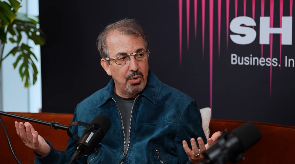 John Hight sitting, arms palm up, in a blue suede jacket and thin glasses. He sits on a red couch, and microphones are blurry in the foreground.