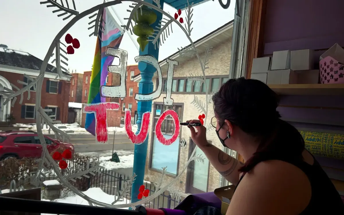 A person wearing glasses and a facemask paints a festive "ICE OUT" design on a shop window. A snowy street can be seen outside.