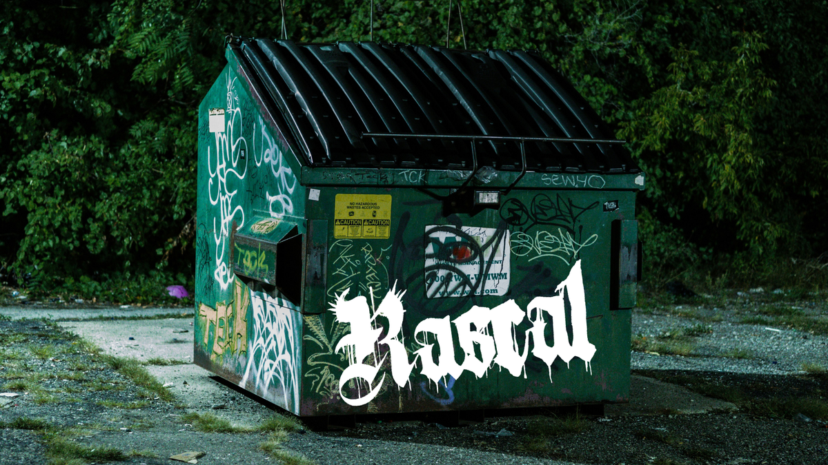 Green dumpster in an urban park at night. Grafitti in dozens of colors cover the exterior, including Rascal's white logo.