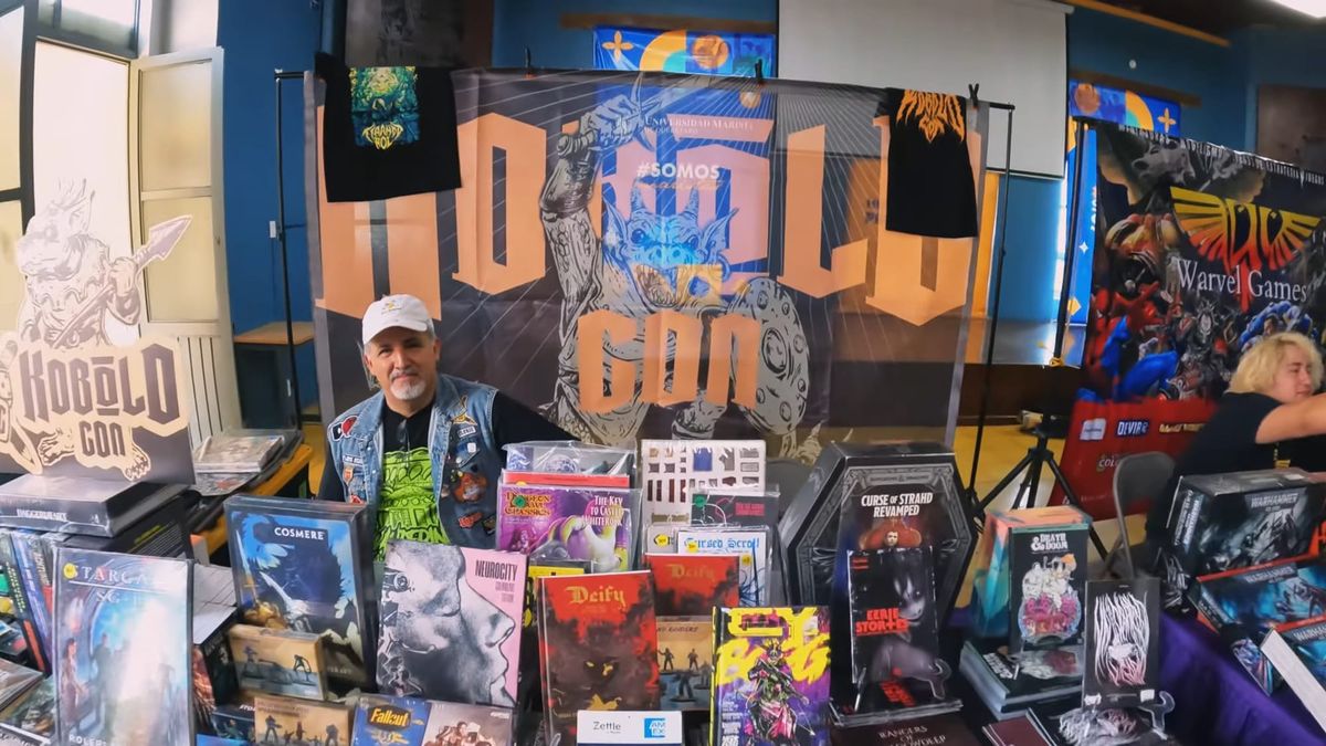 Hugo Hernandez,a mexican man sitting behind a convention table full of games like Cy-Borg, Nuerocity, Deify, Cosmere