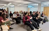 White, well-lit room full of people at a convention, most sitting in black chairs or on a bench. They face one direction and are chatting amongst themselves.