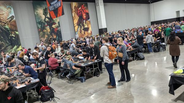 Crowd shot of Adepticon 2025. Tables full of wargame players line a grey-and-white convention hall.