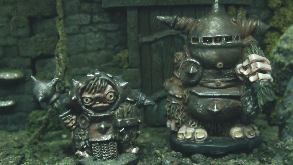 A photograph of two goblin miniatures, Grumple who is armoured and Blombor who is tall and armoured and furry, holding a club. They are standing in front of a wall with moss and a door.