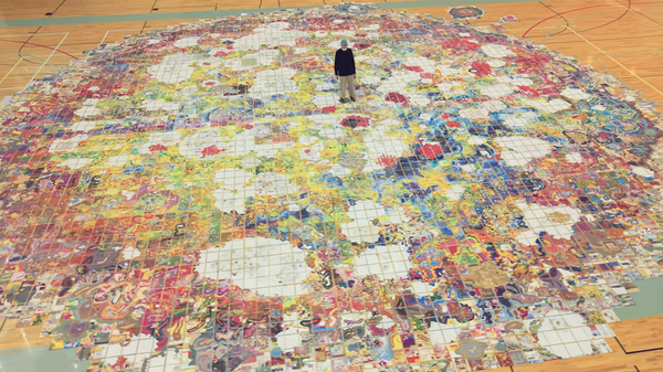 A man in a black sweater and blue cap stands in the middle of a multicolored map spread across the floor of a gymnasium.