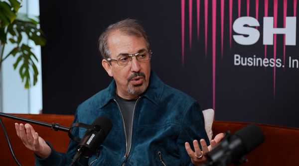 John Hight sitting, arms palm up, in a blue suede jacket and thin glasses. He sits on a red couch, and microphones are blurry in the foreground.