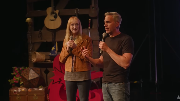 Two people with microphones on a stage decorated with instruments, chests, a big d20 and D&D's brand ampersand.