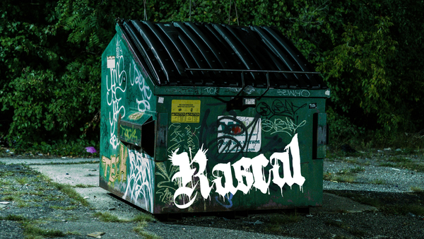 Green dumpster in an urban park at night. Grafitti in dozens of colors cover the exterior, including Rascal's white logo.