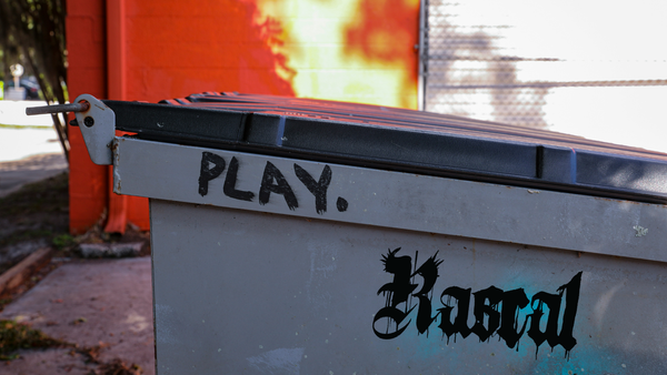 Grey dumpster with black lid in front of an orange building. The worlds "PLAY" and "RASCAL" are spray painted on the side in black.