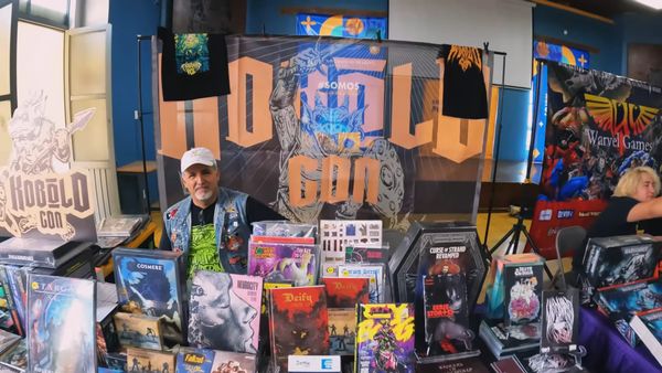 Hugo Hernandez,a mexican man sitting behind a convention table full of games like Cy-Borg, Nuerocity, Deify, Cosmere