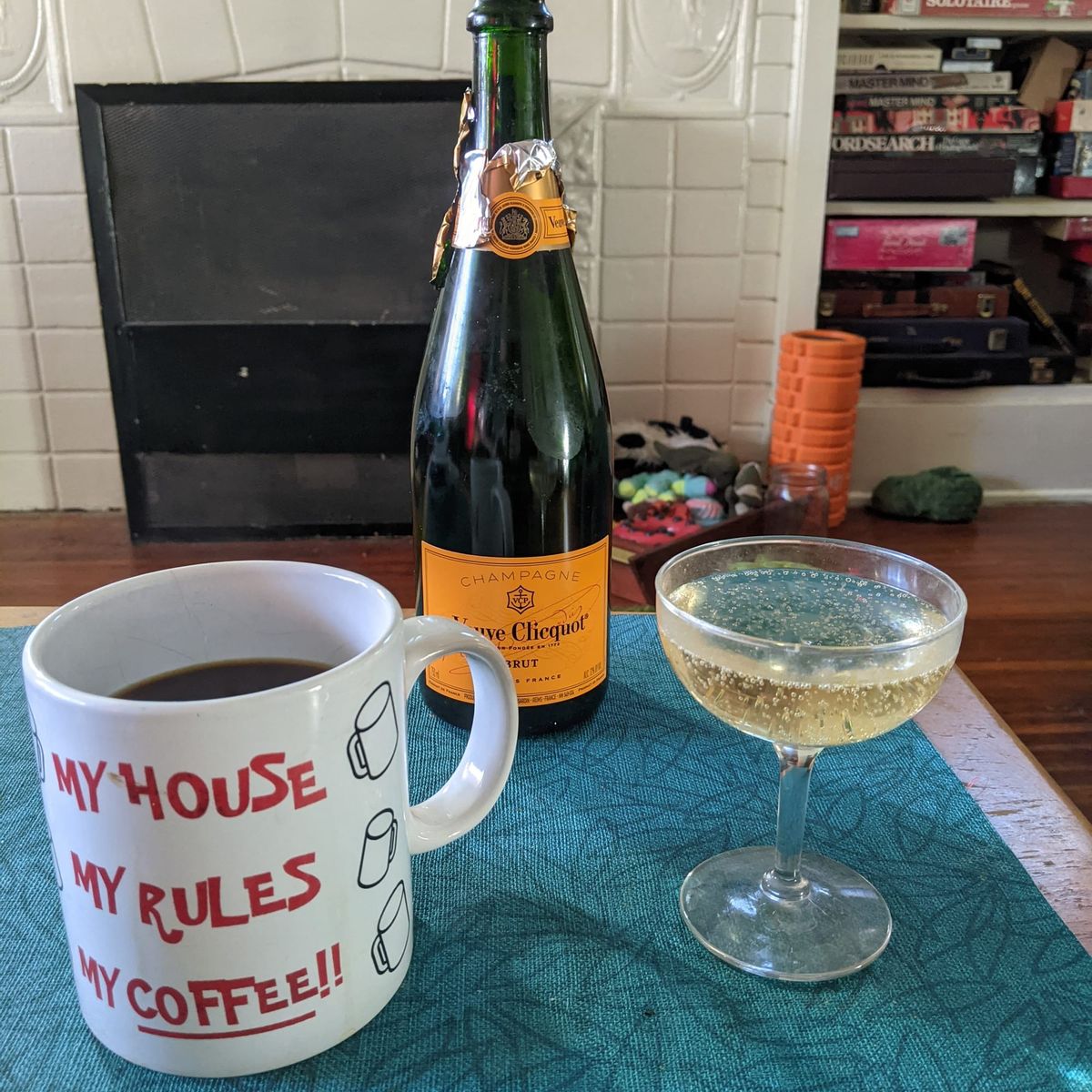 A coffee mug full of coffee, a bottle of Veuve Clicquot champagne, and a coup glass full of bublbling liquid on a coffee table in the morning light