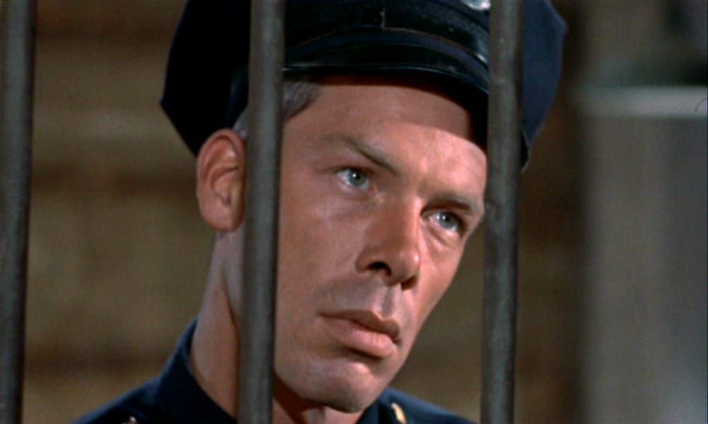 Lee Marvin dressed as a cop looking through the bars of a cage.