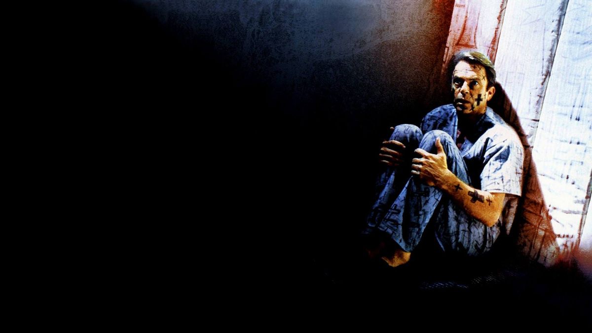 A man covered in black handdrawn crosses wearing hospital clothes and cowering in the corner of a padded cell