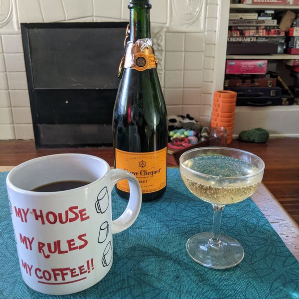 A coffee mug full of coffee, a bottle of Veuve Clicquot champagne, and a coup glass full of bublbling liquid on a coffee table in the morning light