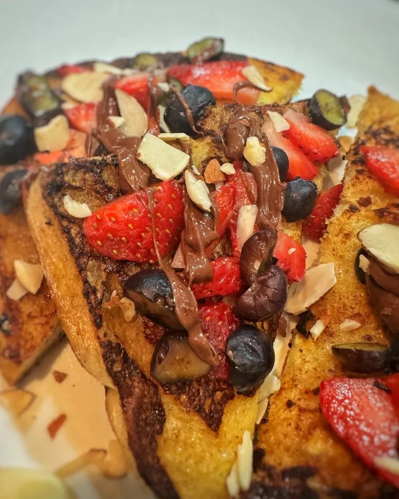 Close-up of sourdough French toast topped with Nutella, fresh strawberries, blueberries, and sliced almonds on a white plate.