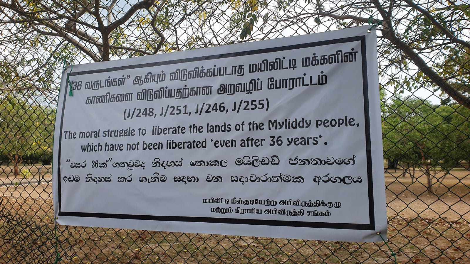 A protest banner displayed outside the military-controlled High Security Zone in Valikamam North calls for the liberation of lands belonging to the people of Myliddy, declaring that even after 36 years, thousands of displaced Tamil families remain denied access to their ancestral homes and farmlands.
