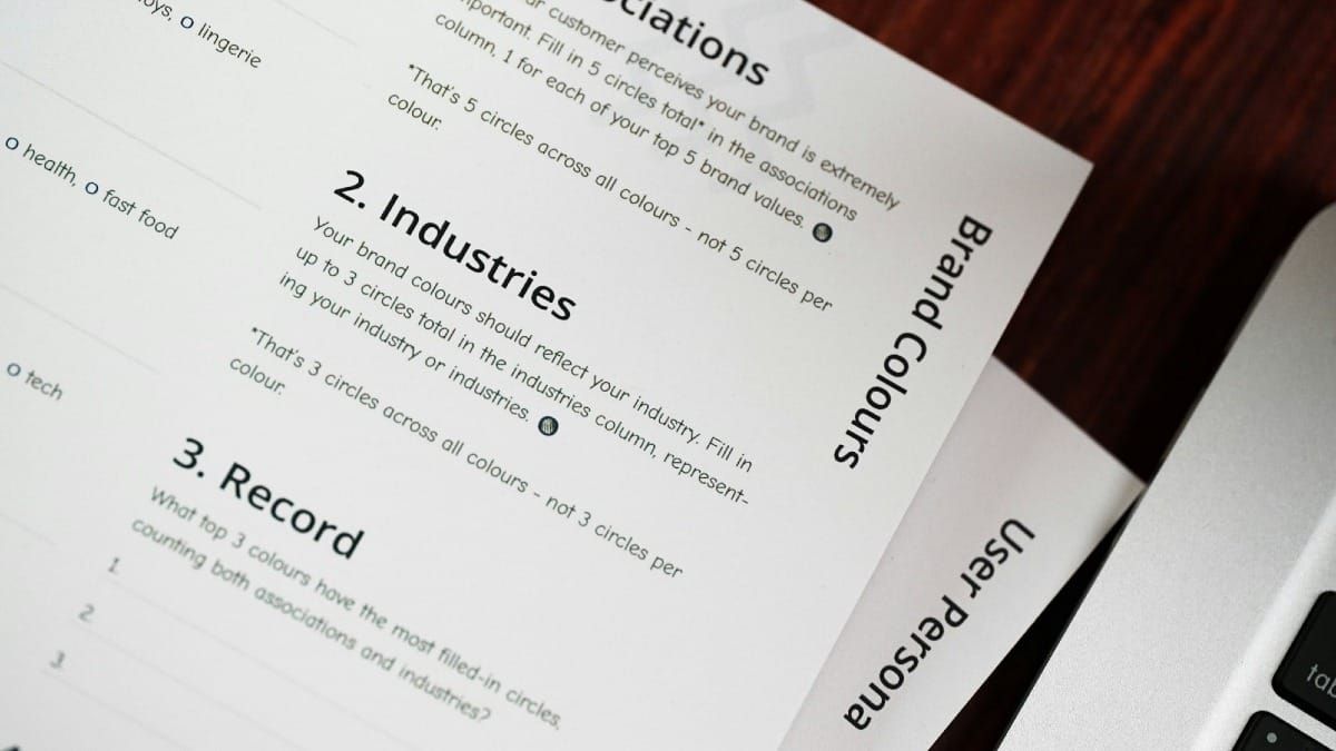 An angled shot of brand development worksheets on a dark desk next to a laptop. Visible headings on the top page include "2. Industries", "3. Record", and "Brand Colours".