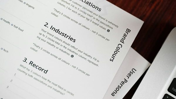 An angled shot of brand development worksheets on a dark desk next to a laptop. Visible headings on the top page include "2. Industries", "3. Record", and "Brand Colours".