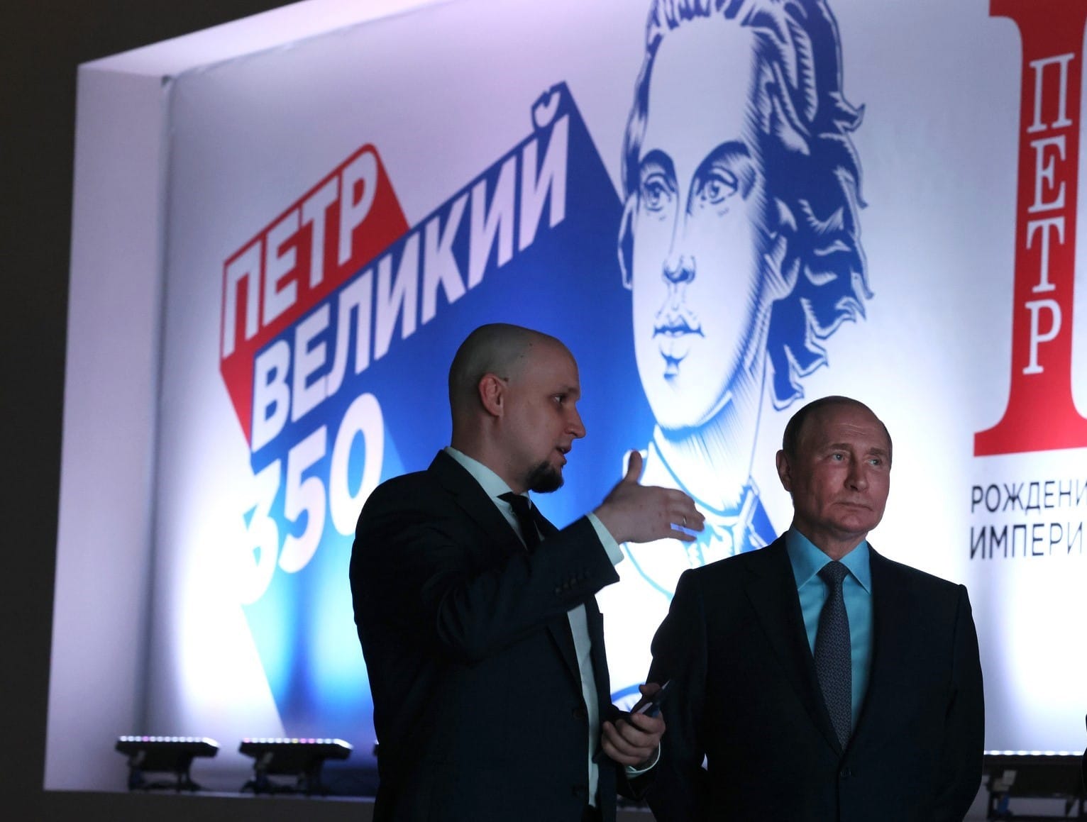 Vladimir Putin standing before an illuminated display devoted to Peter the Great’s 350th anniversary, bearing Peter’s portrait and the Russian words “Birth of the Empire.”