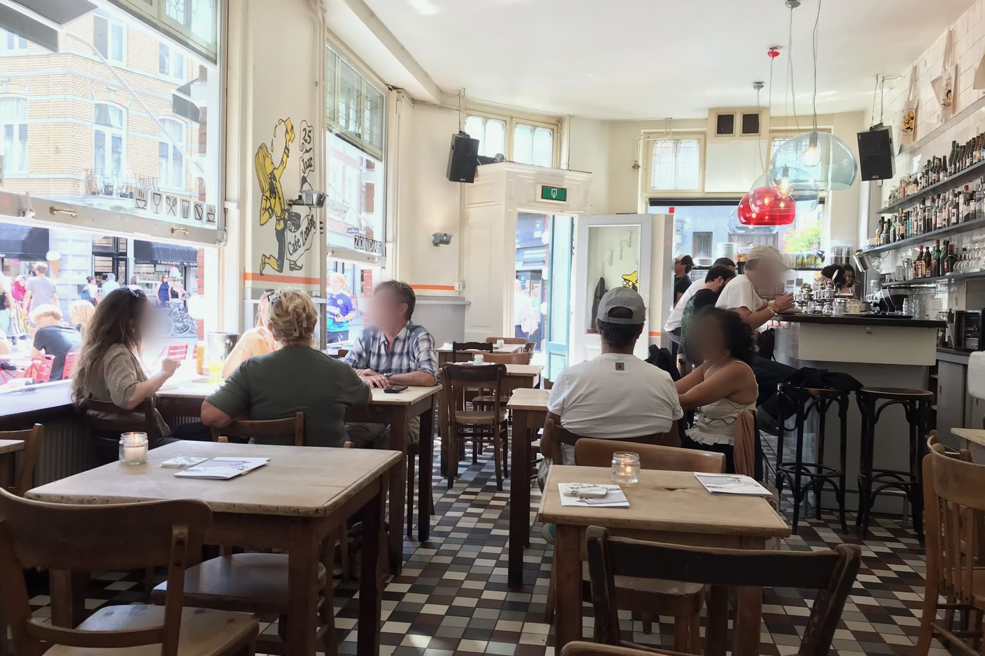 Het interieur van Café Zondag, met bezoekers in gesprek aan tafels bij de open ramen en de bar.