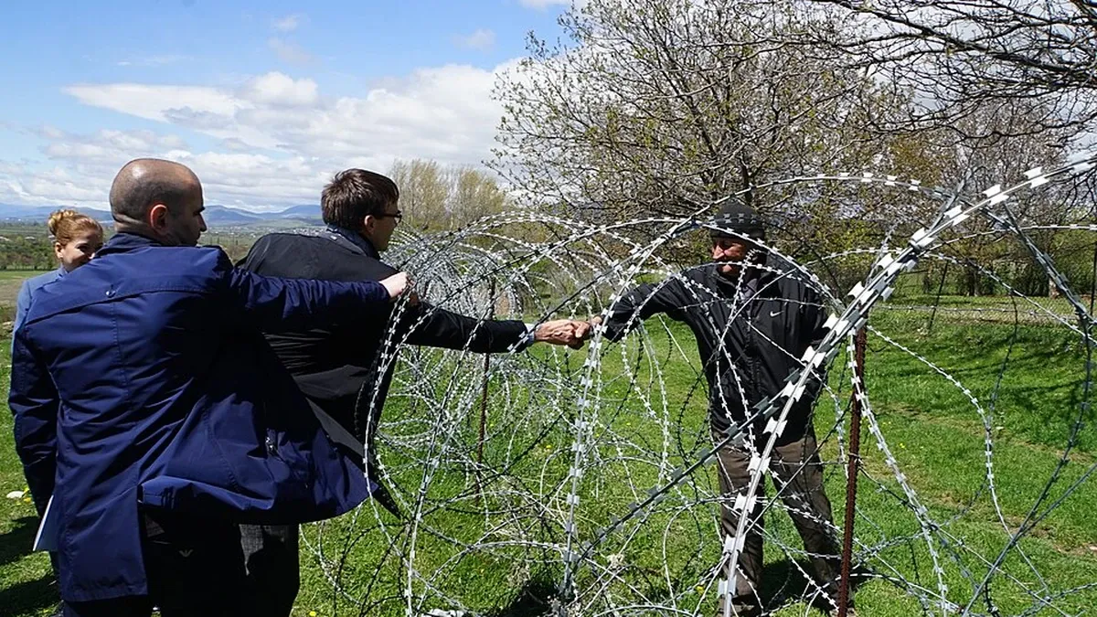 Residents on opposite sides of the boundary line near South Ossetia in Georgia reach toward one another through coils of razor wire in a rural landscape.