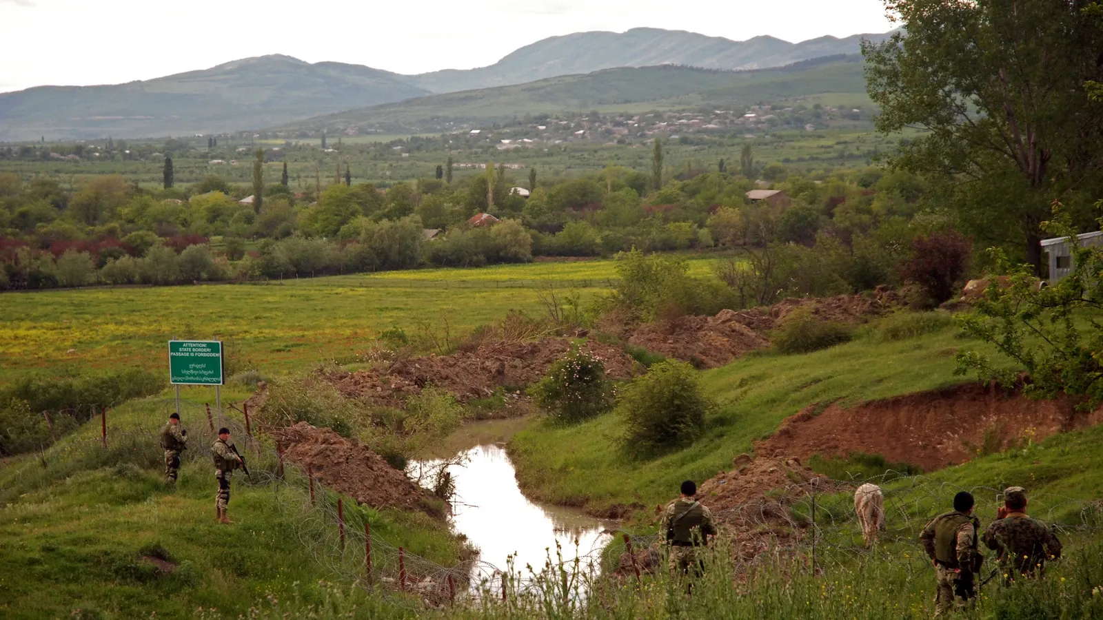 Rural landscape near the South Ossetia boundary in Georgia, with soldiers, fencing and a trench cutting through fields beneath hills in the background.