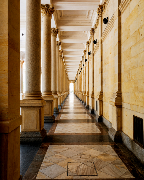A long stone gallery with repeated columns and arches leading towards a sunlit courtyard.