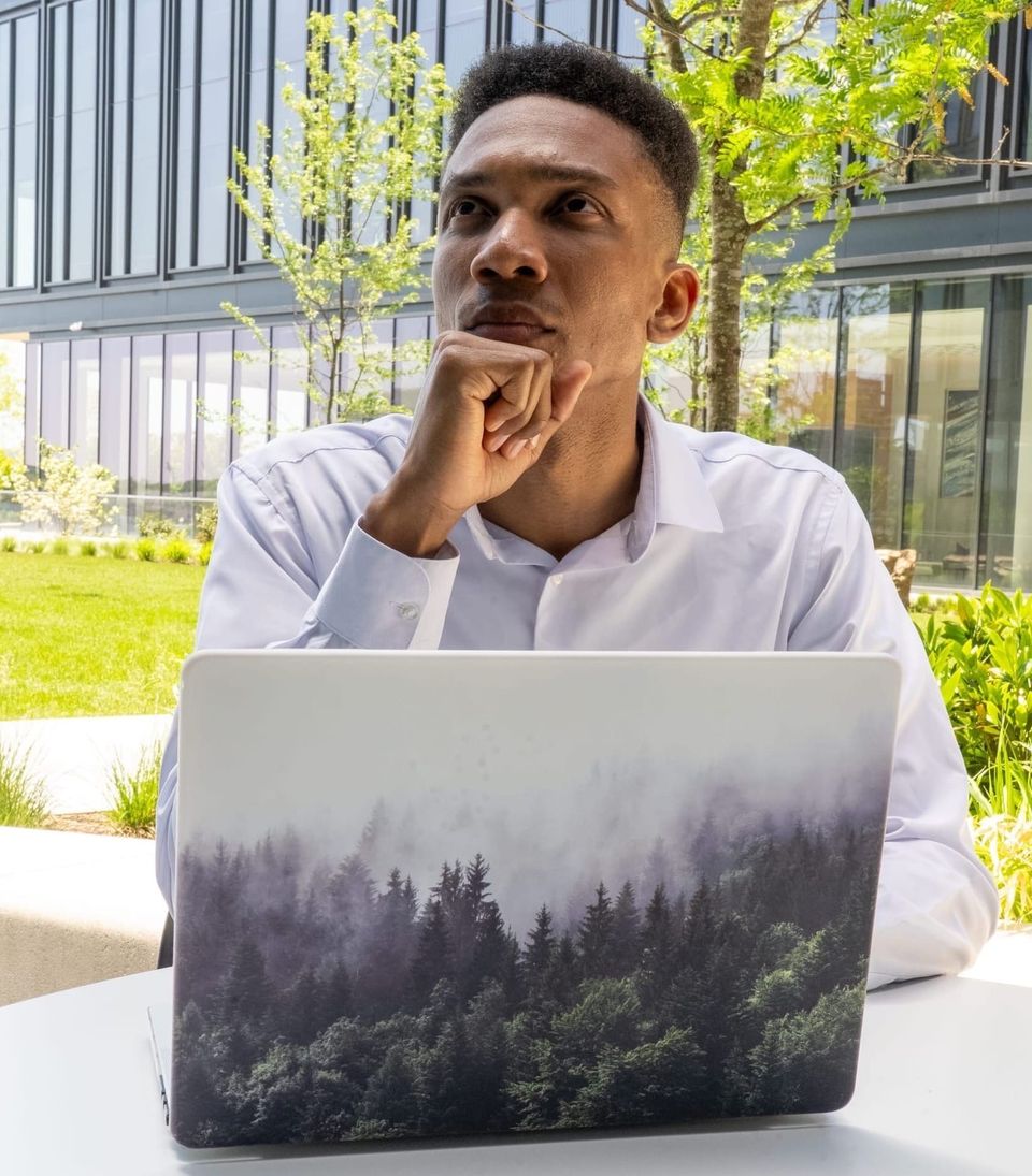 The Author is sitting down at a table, pensively looking off into the distance using a laptop computer.
