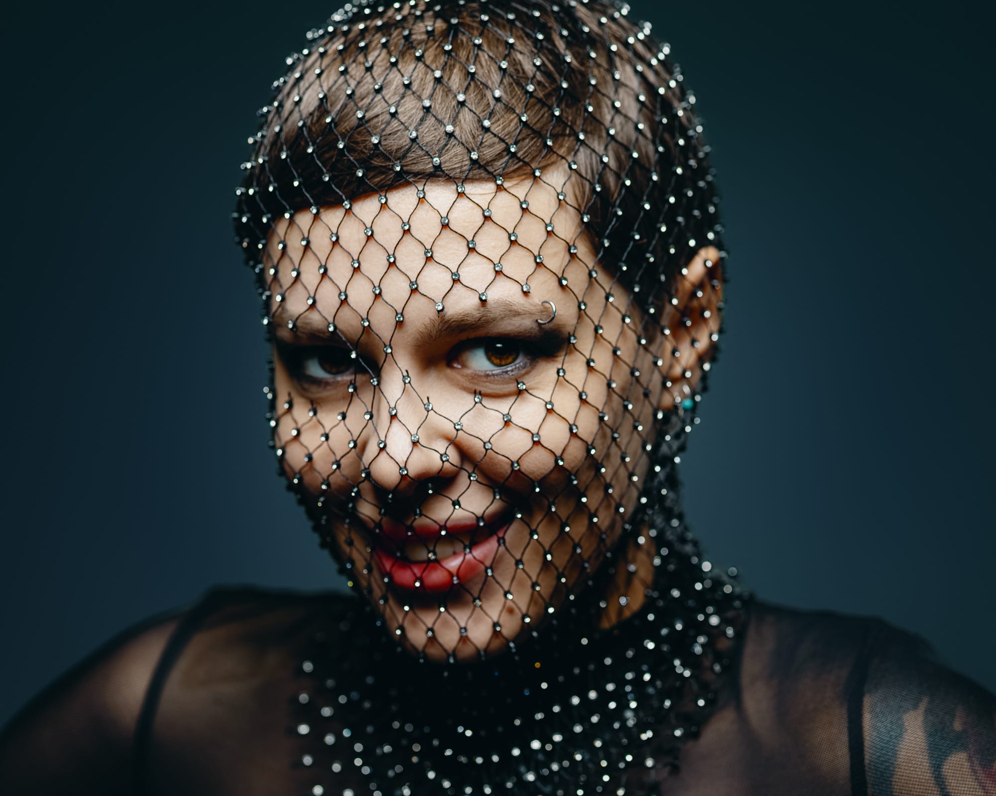 Close-up portrait through a beaded mesh veil, red lipstick, intense eye contact, dark studio background.