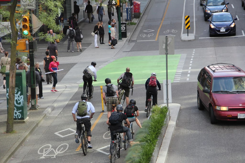 Seven cyclists ride down a protected bike lane in downtown Vancouver. Pedestrians are on the sidewalk, and cars drive down the street.